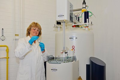 Eine Frau im weißen Chemie-Kittel steht vor einem Forschungsgroßgerät.