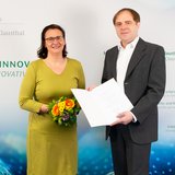 Die Universitätspräsidentin und ein neu ernannter Professor stehen vor einer Wand mit einem Logo der TU Clausthal.