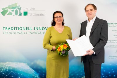 Die Universitätspräsidentin und ein neu ernannter Professor stehen vor einer Wand mit einem Logo der TU Clausthal.