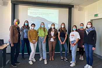 Studierende tragen Masken und stehen vor einem Projektorbildschirm 