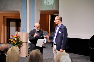 Ein Mann erhält von einem anderen eine Urkunde in einem festlichen Rahmen.