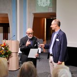 Ein Mann erhält von einem anderen eine Urkunde in einem festlichen Rahmen.