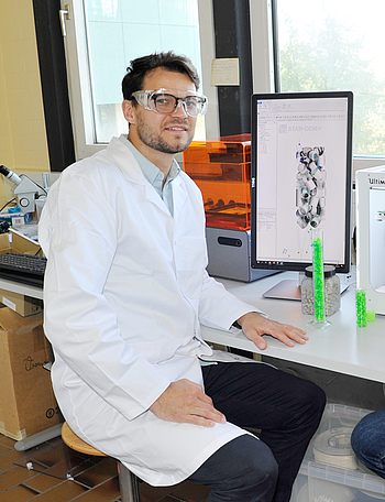 Eine Person mit Laborkittel und Schutzbrille sitzt vor einem Monitor 