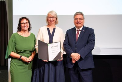 Drei Personen stehen festlich gekleidet nebeneinander, die Frau in der Mitte hält eine Urkunde in der Hand.