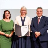 Drei Personen stehen festlich gekleidet nebeneinander, die Frau in der Mitte hält eine Urkunde in der Hand.