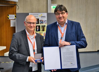 Zwei Männer, einer mit einer Medaille, der andere mit einer Urkunde, stehen in einem Hörsaal.