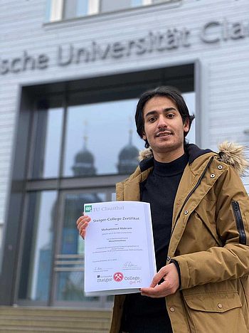 Eine Person mit einem Steiger-College-Zertifikat vor dem Eingang zum Hauptgebäude der TU Clausthal
