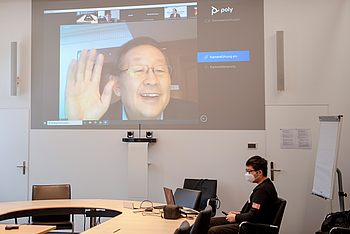 Eine Person sitzt vor einer großen Projektionsfläche, die eine Online-Videokonferenz zeigt