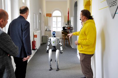 Ein Roboter marschiert durch einen Gang und wird von drei Personen betrachtet.  