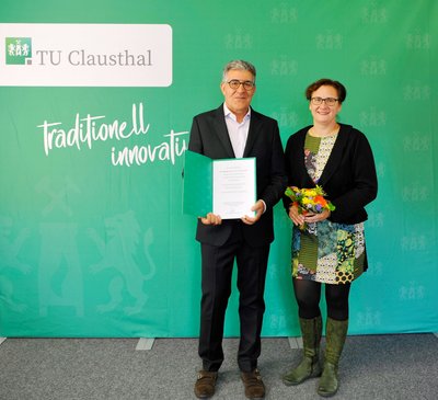 Ein Mann mit Urkunde und eine Frau mit Blumenstrauß stehen vor einer grünen Wand.