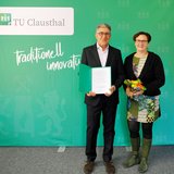 Ein Mann mit Urkunde und eine Frau mit Blumenstrauß stehen vor einer grünen Wand.