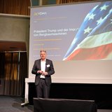 Ein männlicher Referent steht vor eine Leinwand, auf der eine USA-Flagge zu sehen ist. 