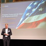 Ein männlicher Referent steht vor eine Leinwand, auf der eine USA-Flagge zu sehen ist. 