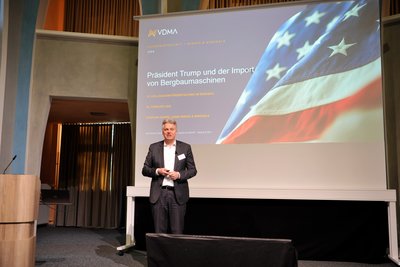 Ein männlicher Referent steht vor eine Leinwand, auf der eine USA-Flagge zu sehen ist. 