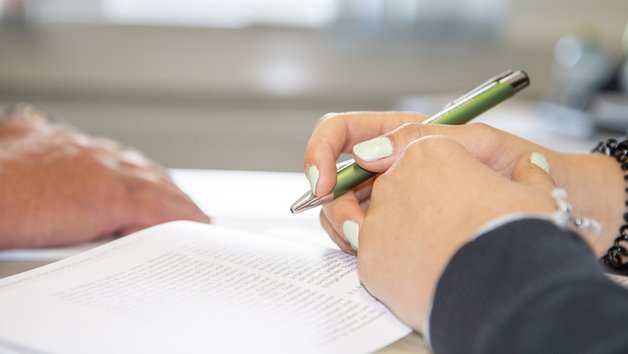Vier Händ die auf einem Tisch liegen. Eine Person hält einen Stift in der Hand