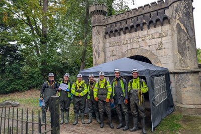 Eine Gruppe in Bergbaukleidung steht vor einem Stolleneingang. 