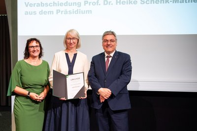 Drei Personen stehen festlich gekleidet nebeneinander, die Frau in der Mitte hält eine Urkunde in der Hand.