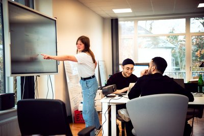 In einem innovativen Lernumfeld erklärt eine junge Frau zwei jungen Männern am Bildschrim etwas.