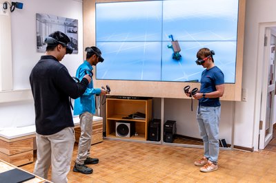 Drei Studierende stehen mit VR-Brillen vor einer großen Leinwand. 
