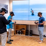 Drei Studierende stehen mit VR-Brillen vor einer großen Leinwand. 