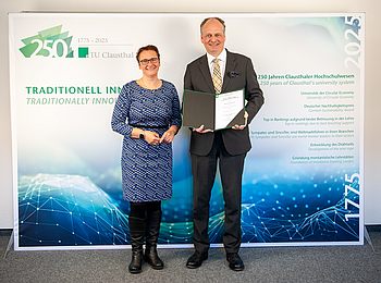 Sylvia Schattauer und Matthias von Kaler stehen gemeinsam vor der TU-Foto-Wand mit der aufgeklappten Urkunde.