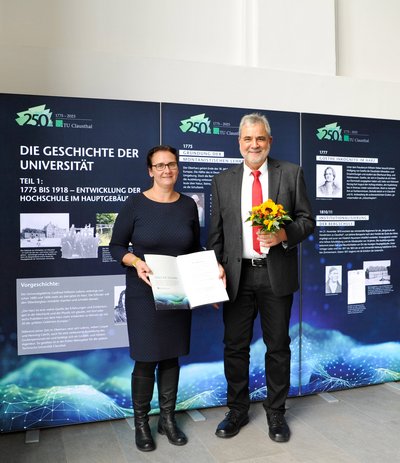 Eine Frau mit einer Urkunde in der Hand und ein Mann mit Blumen stehen vor einer Ausstellungswand.