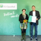 Eine Frau und ein Mann mit einer Urkunde stehen vor einer grünen Wand.