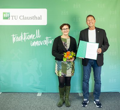 Eine Frau und ein Mann mit einer Urkunde stehen vor einer grünen Wand.