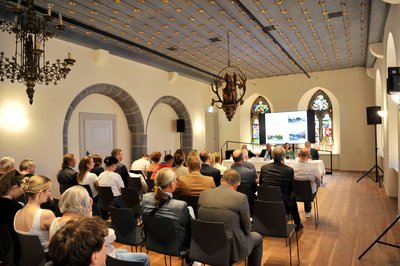 Zahlreiche Menschen sitzen in einem Saal eines historischen Gebäudes. 